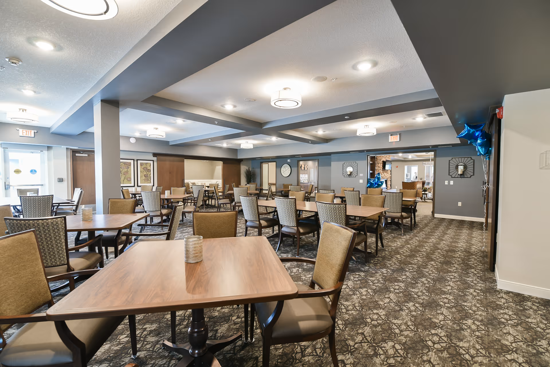 A spacious dining room in a senior living facility with multiple wooden tables and cushioned chairs arranged neatly. The room has patterned carpet flooring, gray walls, and ceiling lights. Blue star-shaped balloons are tied to a wall on the right side. There are exit signs above doors at the far end of the room.