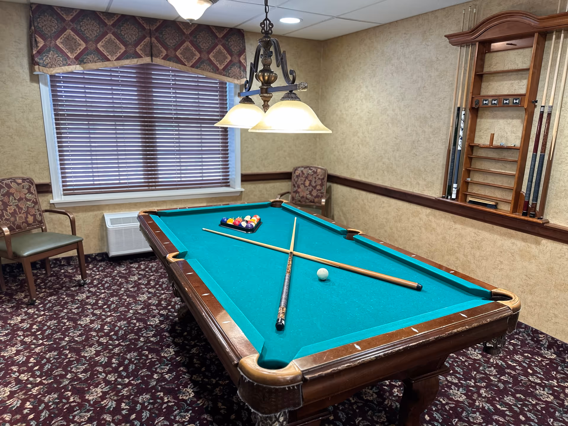 A billiards room with a pool table in the center, two pool cues crossed on the table, a rack of billiard balls, two floral-patterned chairs, a window with closed blinds and a valance, and a wall-mounted wooden cue rack holding additional pool cues.