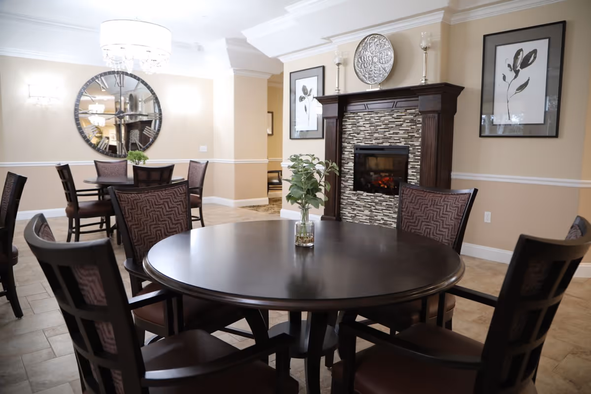 Interior view of a dining area with round dark wood tables and matching chairs. A small vase with green foliage is placed on the nearest table. The room features beige walls with white trim, a decorative fireplace with a tiled surround, and framed botanical artwork on the walls. A large round mirror and a chandelier are visible in the background.