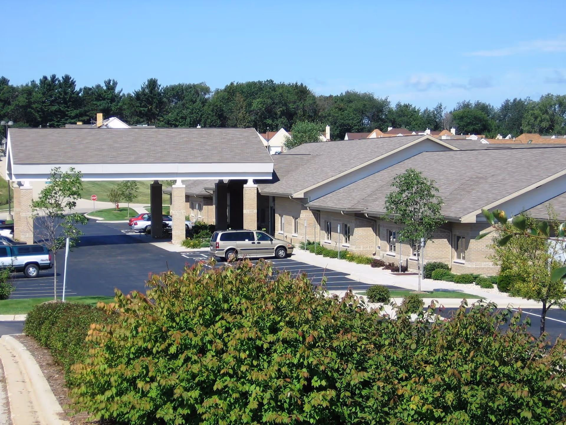 Exterior view of Coventry Village senior living facility showing a single-story building with a covered entrance, parking lot with several cars, and landscaped greenery in the foreground under a clear blue sky.