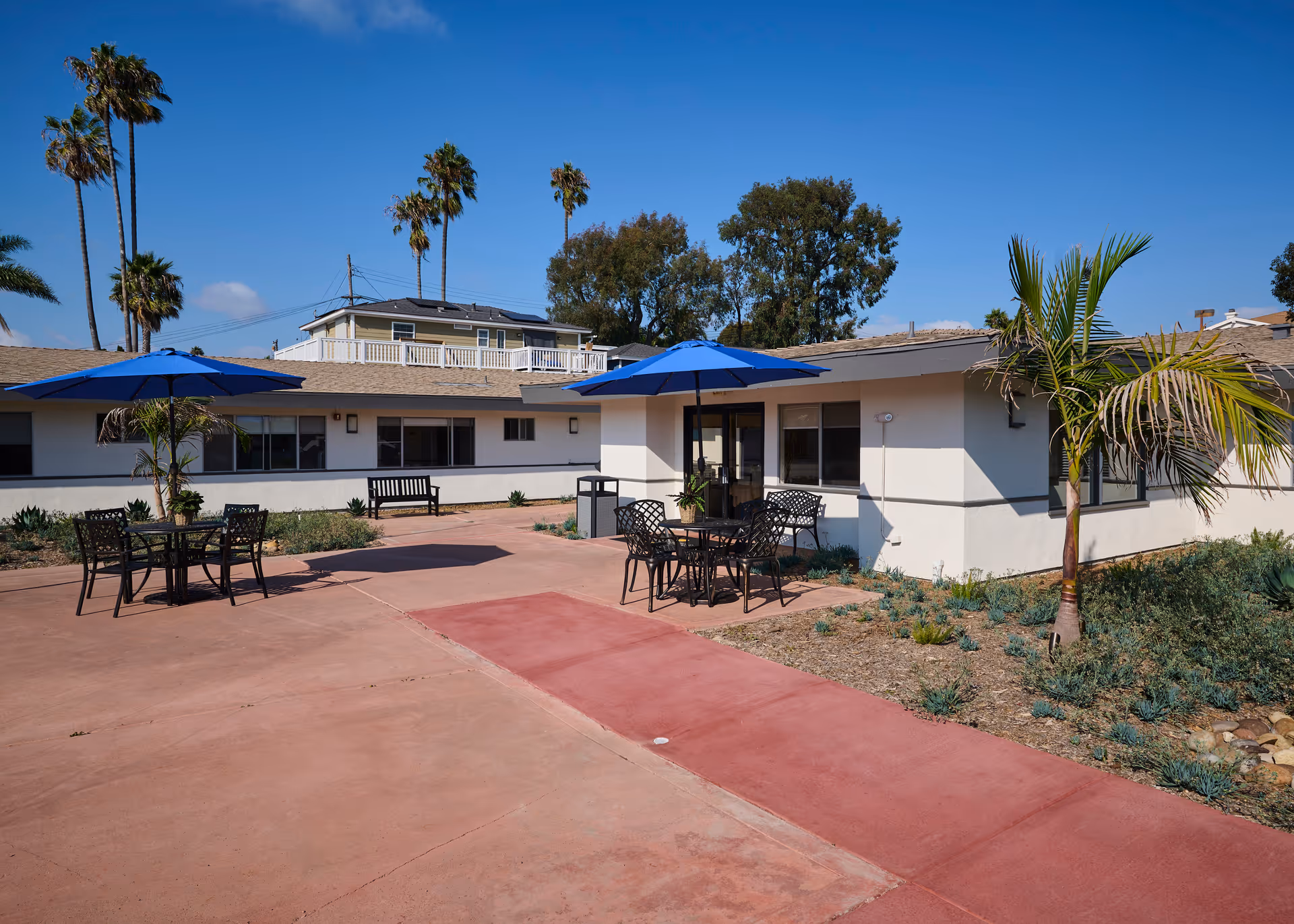 Outdoor patio area at Encinitas Nursing and Rehabilitation Center with tables and chairs under blue umbrellas, surrounded by palm trees and landscaping, adjacent to a single-story building under a clear blue sky.