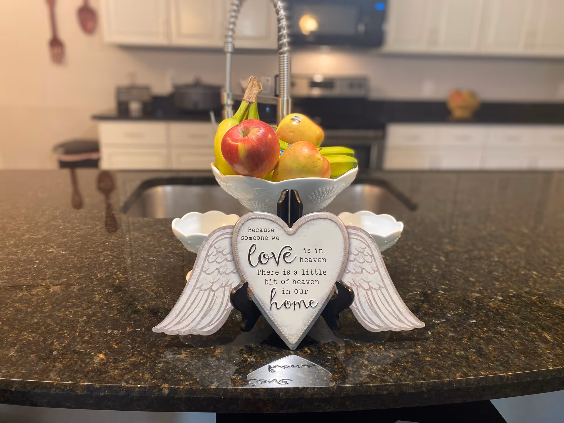A kitchen countertop with a decorative heart-shaped sign with wings that reads 'Because someone we love is in heaven there is a little bit of heaven in our home'. Behind the sign is a white bowl filled with bananas, apples, and pears. The background shows a kitchen sink, stove, and white cabinets.