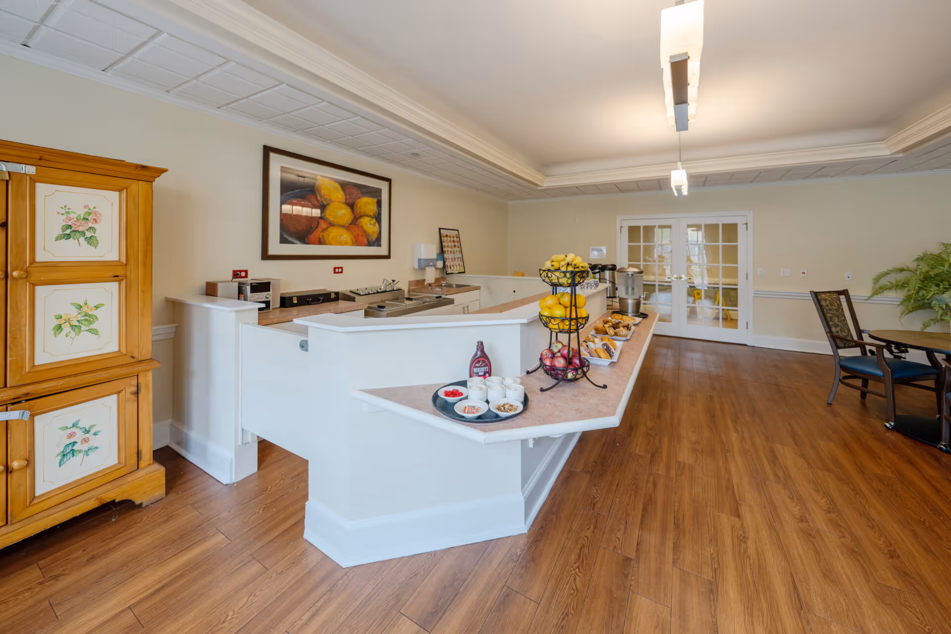 Interior dining and serving area with a buffet counter stocked with fruit and pastries, wooden flooring, and seating.