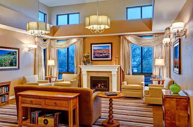 Well-lit communal living room with sofas and armchairs arranged around a fireplace beneath large windows and chandeliers.