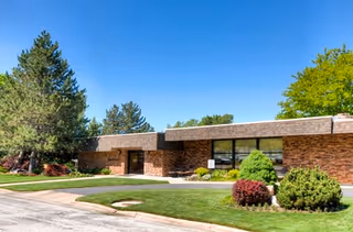 Exterior view of a single-story brick building with a flat roof, surrounded by well-maintained green lawns, shrubs, and trees under a clear blue sky.