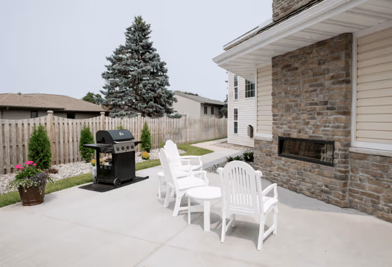 Outdoor patio area with three white chairs and a small round table on a concrete surface next to a stone wall with a built-in fireplace. A black grill is positioned nearby, and there is a wooden fence and some greenery in the background.