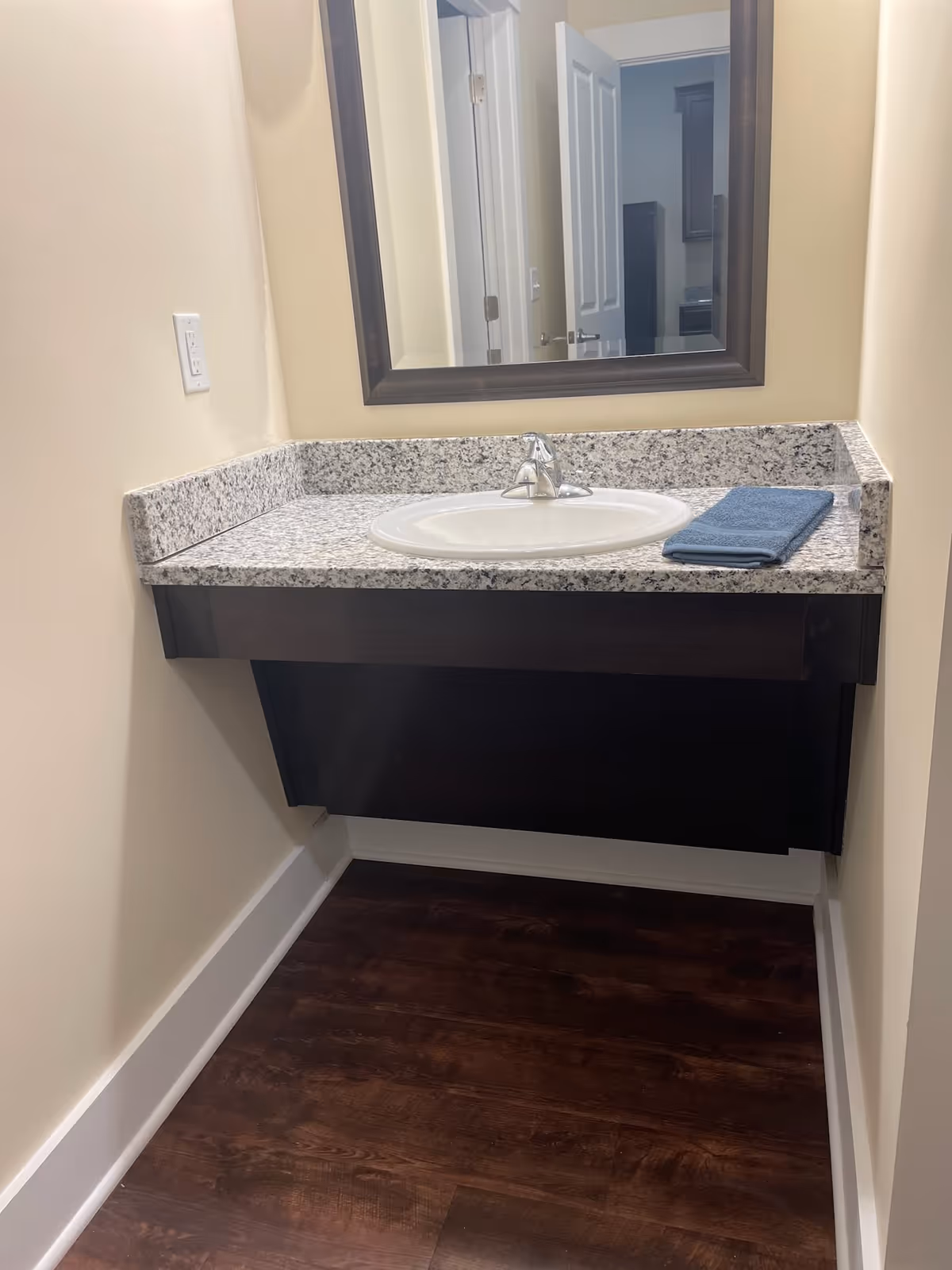 A bathroom vanity with a granite countertop, an oval sink, a silver faucet, and a folded blue towel on the right side. Above the vanity is a large rectangular mirror with a dark wooden frame. The floor is dark wood, and the walls are painted a light beige color.