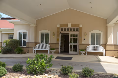 Entrance of a senior living facility with a covered driveway, two white benches on either side of the glass double doors, decorative plants in pots, and some landscaping with bushes and rocks in front.