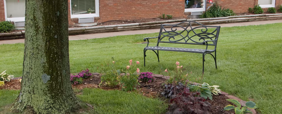 Wrought-iron bench on a grassy lawn beside a flowerbed and tree in front of a brick building.