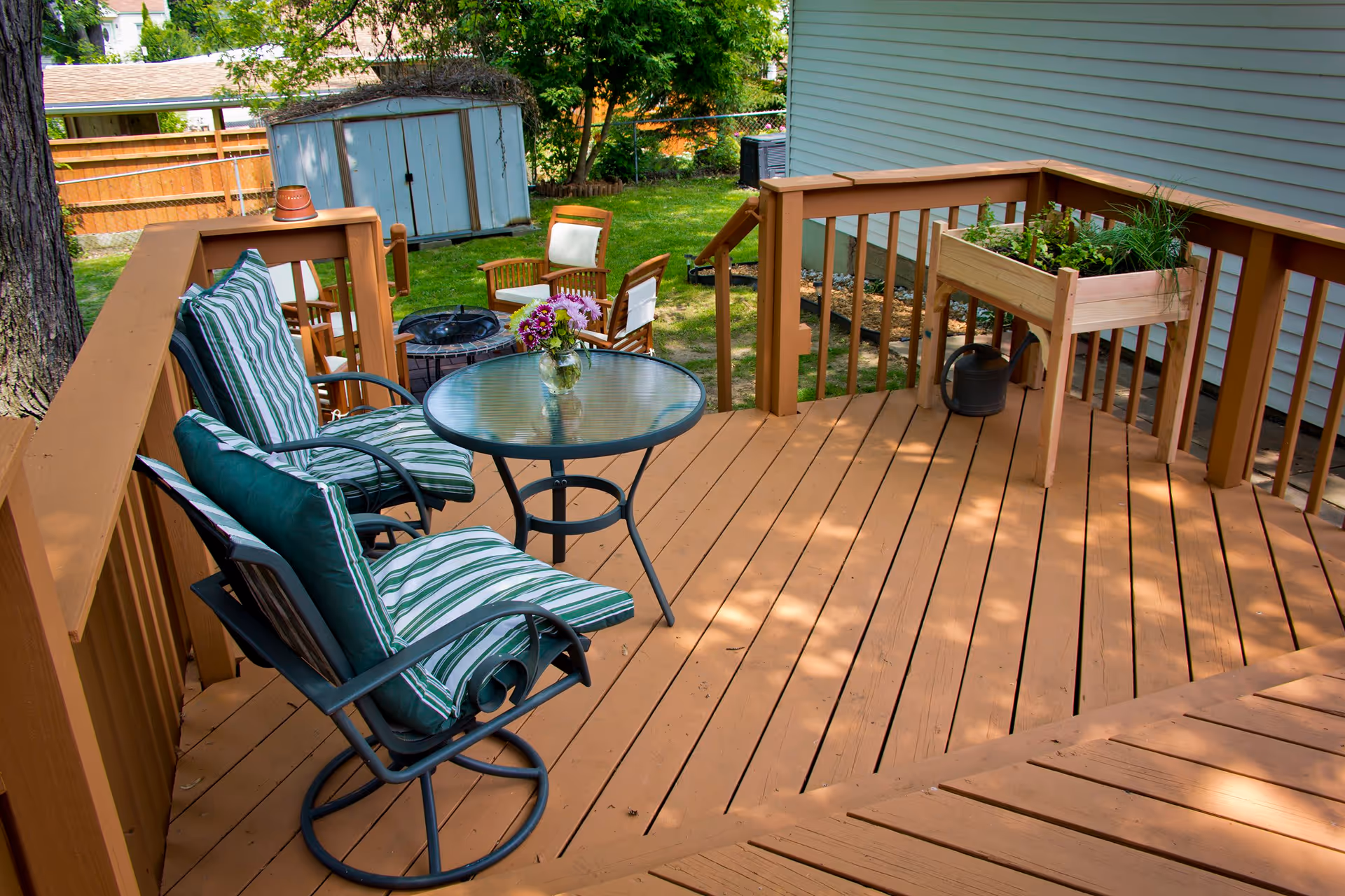 A wooden outdoor deck with two cushioned chairs and a glass-top round table with a vase of flowers. In the background, there are additional wooden chairs around a fire pit, a small garden planter with plants, a watering can, and a shed surrounded by a fenced yard with trees and grass.