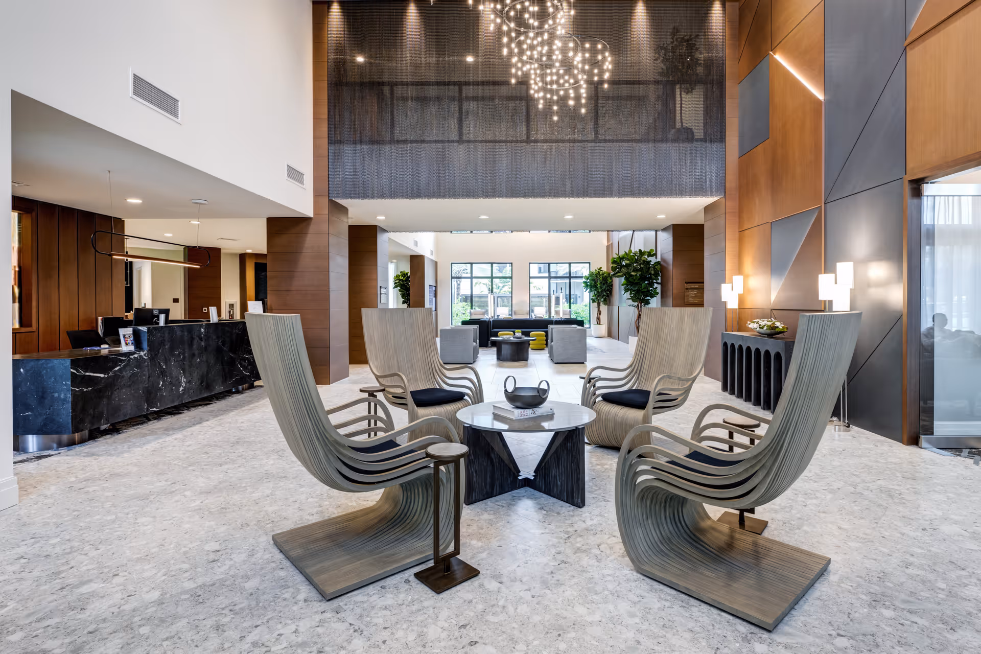 Modern senior living facility lobby with a high ceiling, featuring a circular arrangement of six uniquely designed wooden chairs around a small round table. The space includes a black marble reception desk on the left, large windows at the back letting in natural light, contemporary lighting fixtures, and wood-paneled walls with geometric accents.