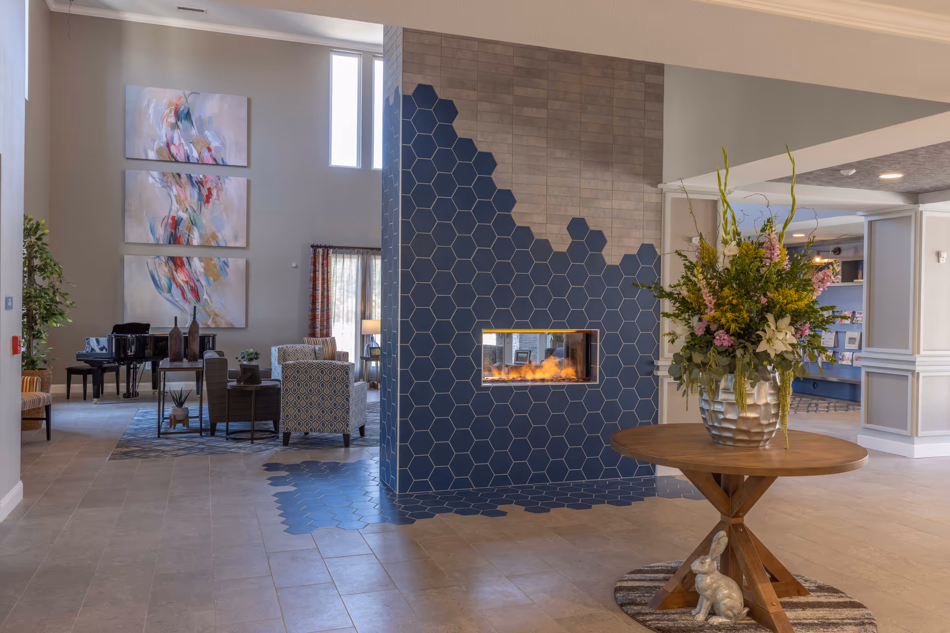 Bright lobby interior with a hex-tiled double-sided fireplace, round table with flowers, and a seating area with piano and large artwork.