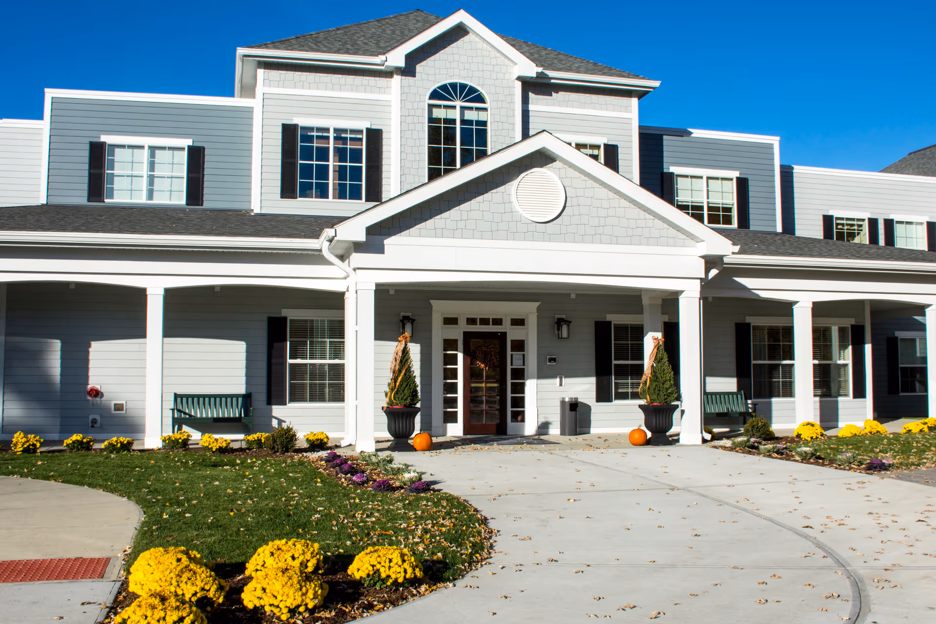 Front exterior view of a senior living facility building with gray siding, white trim, and black shutters. The entrance has a covered porch with two potted plants and pumpkins on either side of the door. There are benches on the porch and yellow flowers planted along the walkway and lawn.