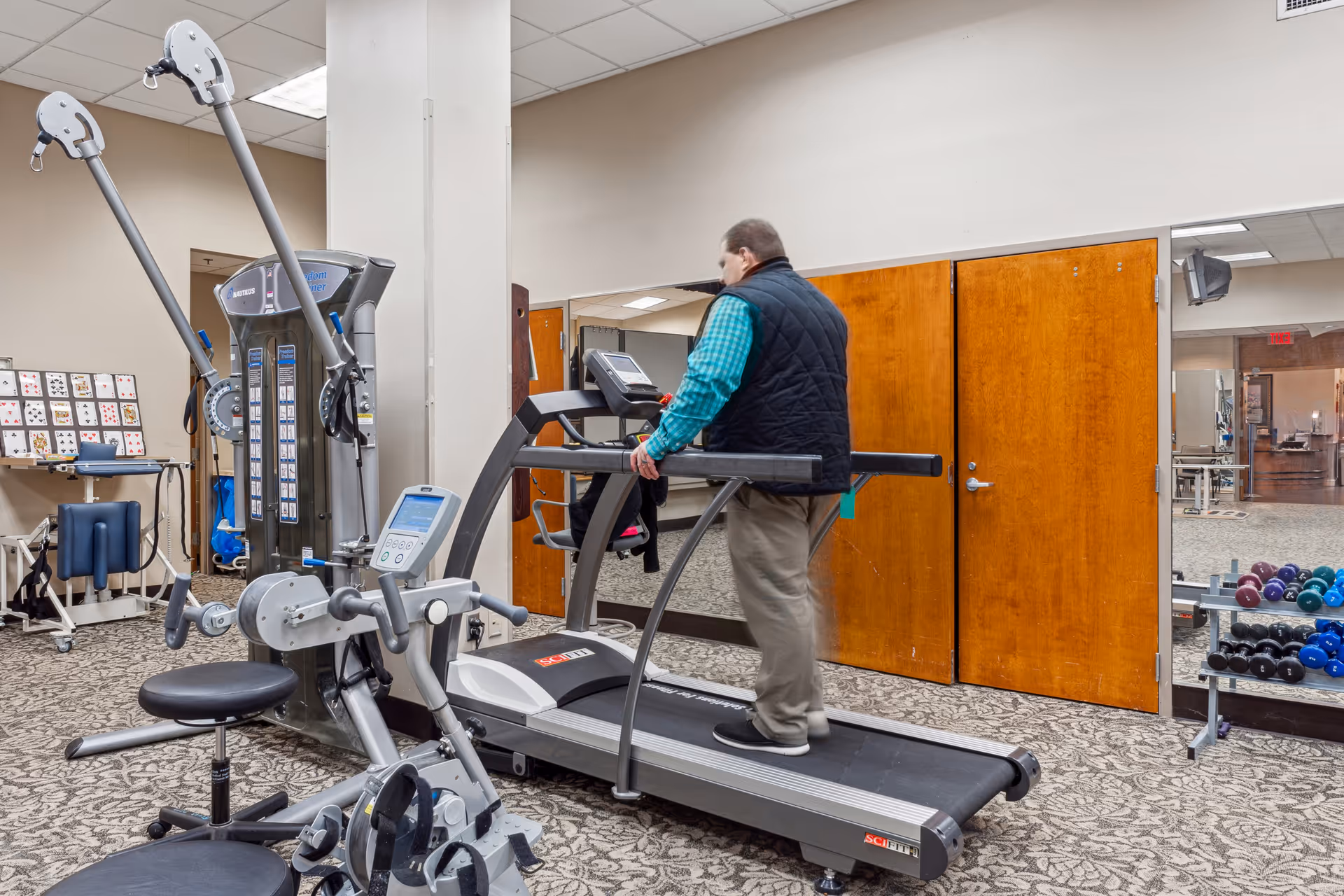 A man walking on a treadmill inside a fitness room with exercise machines and a rack of dumbbells.