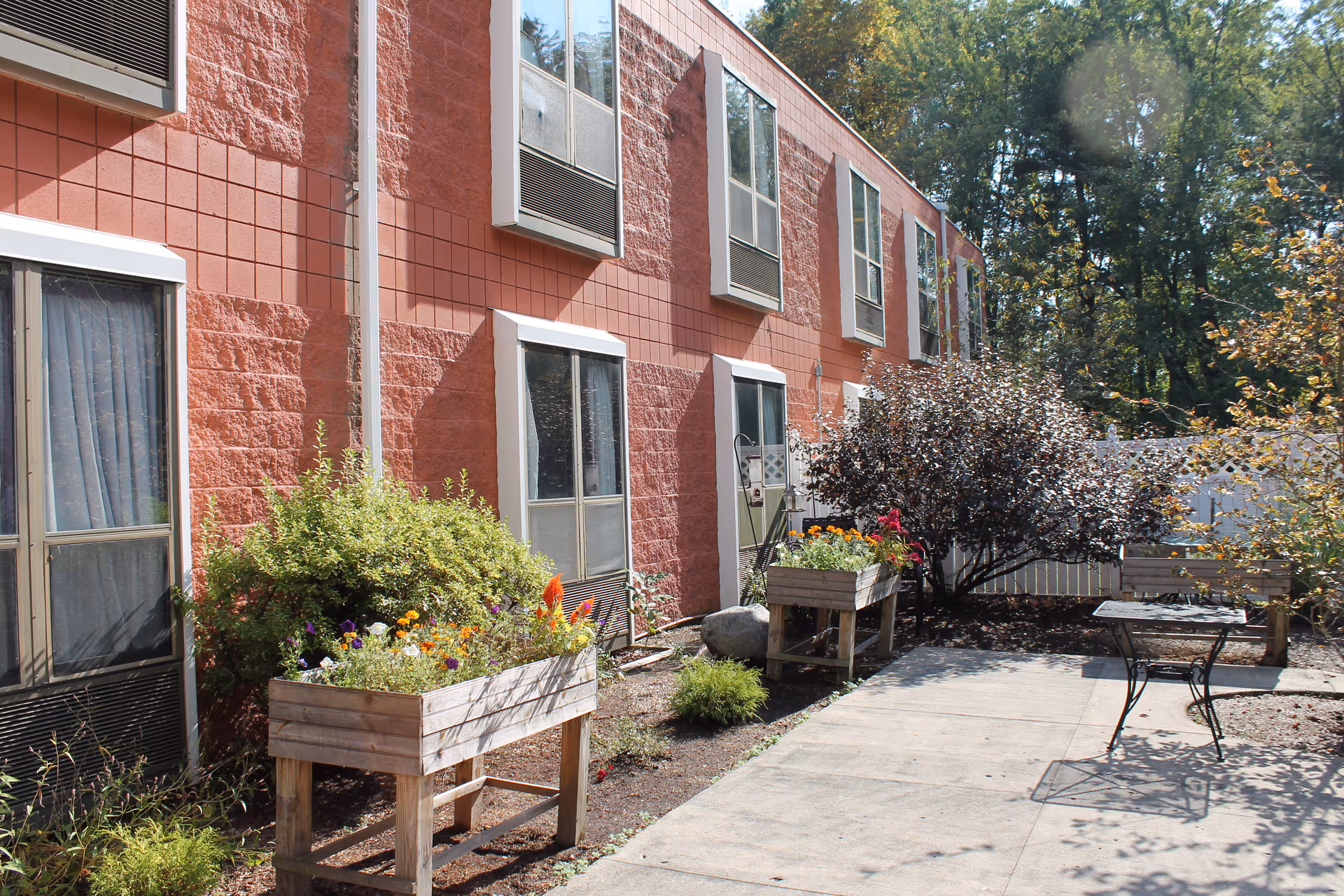Outdoor courtyard area of a building with red brick walls and several windows. The courtyard features raised wooden planters with colorful flowers, a small metal table, and a bench surrounded by trees and shrubs.