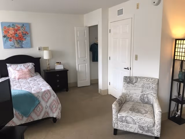 A cozy bedroom with a single bed covered in a patterned quilt and pillows. Next to the bed is a black nightstand with a lamp and some items on it. A colorful floral painting hangs on the white wall above the bed. There is a patterned armchair with seashell designs near a tall black floor lamp with a square lampshade. Two white doors are visible, one slightly open showing a closet with hanging clothes.