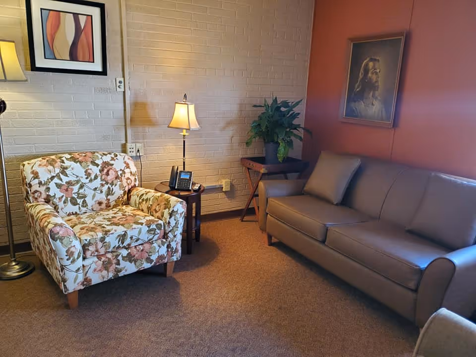 Cozy living room with a floral armchair, leather sofa, side tables with lamps, a potted plant and wall art.