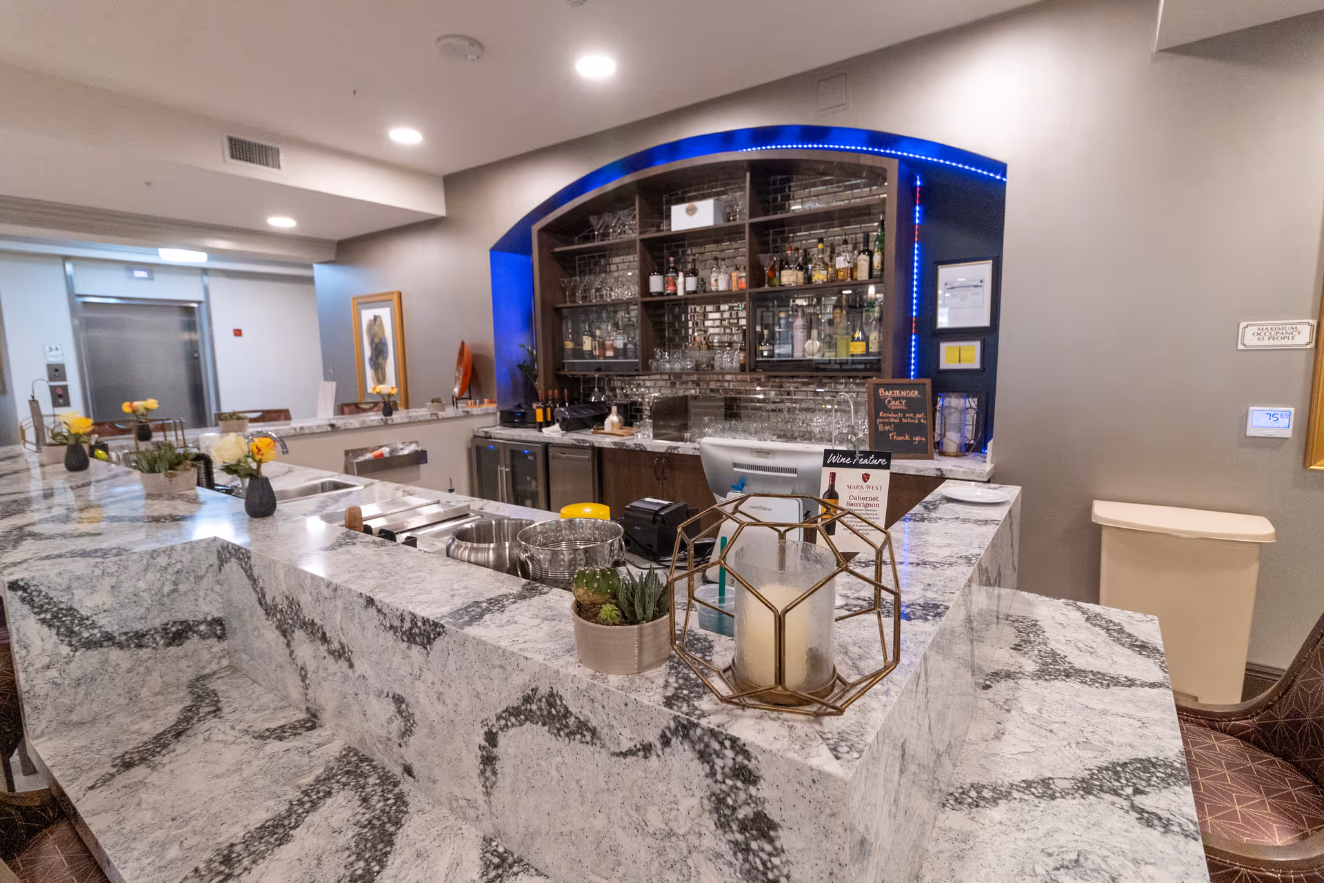 Interior bar and service counter with marble countertops, decorative plants and candle, and shelves of glassware and liquor in the background.