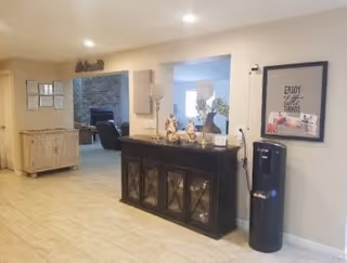 Interior view of a senior living facility reception or common area with a dark wooden cabinet decorated with small figurines and a water dispenser next to it. The wall has a framed picture with the text 'ENJOY the little THINGS'. In the background, there is a stone fireplace and a seating area with chairs. The floor is tiled and the walls are painted light beige.