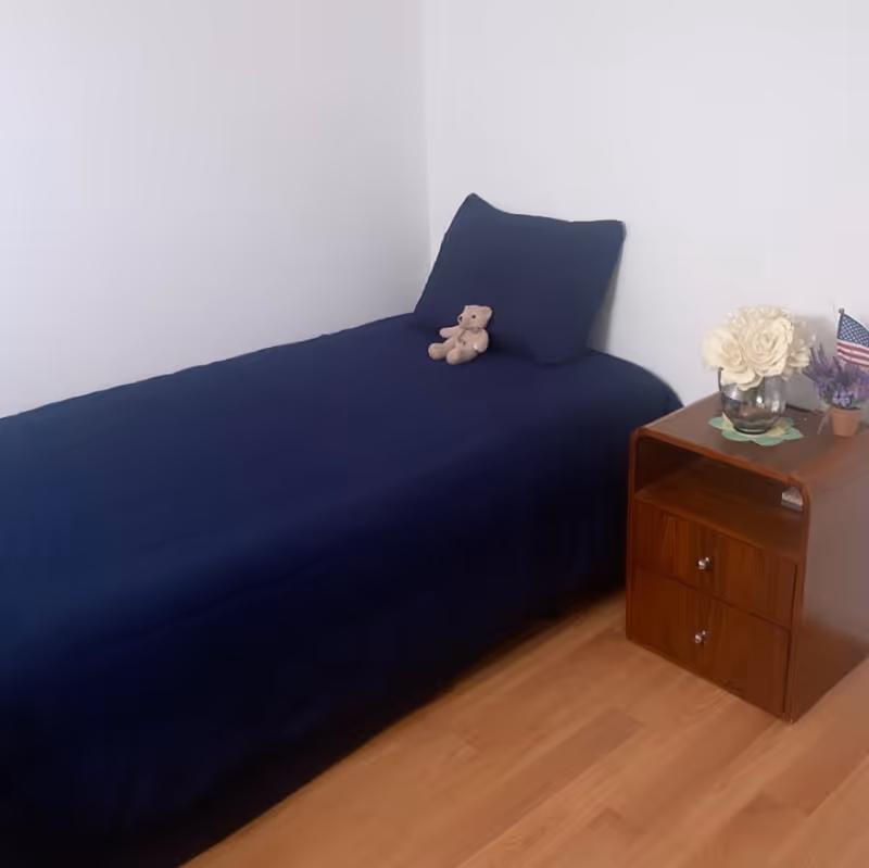 Simple bedroom with a single bed dressed in navy bedding, a small teddy on the pillow, and a wooden nightstand with flowers and a small American flag.