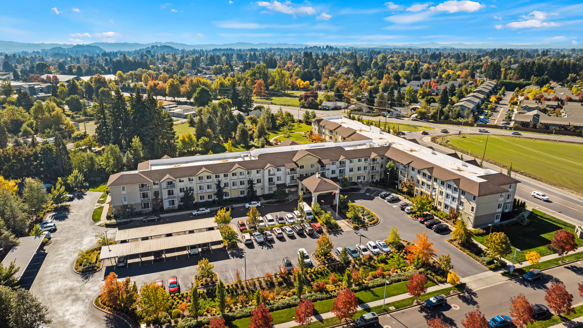 Aerial view of Volante Senior Living of Eugene, showing a large three-story building with a brown roof surrounded by landscaped gardens, parking lots with cars, and trees with autumn foliage under a blue sky with scattered clouds.