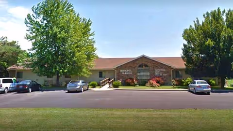 Exterior front view of Bristol Manor Warrensburg, a single-story brick facility with parked cars outside.