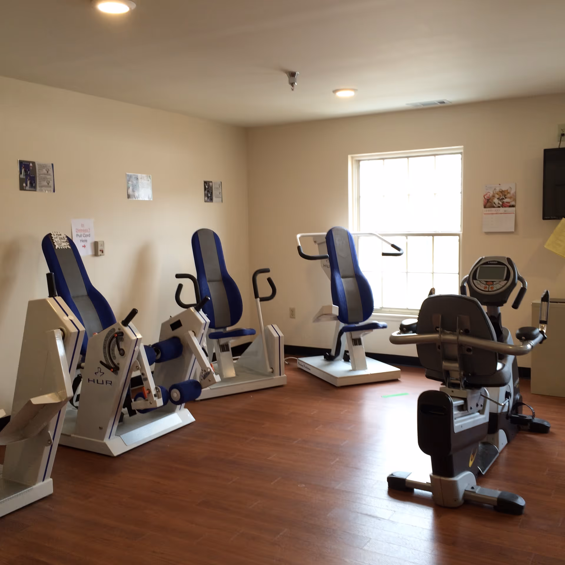 A small exercise room with several pieces of fitness equipment including seated leg press machines and a recumbent stationary bike. The room has wooden flooring, beige walls, a window letting in natural light, and a wall-mounted TV.