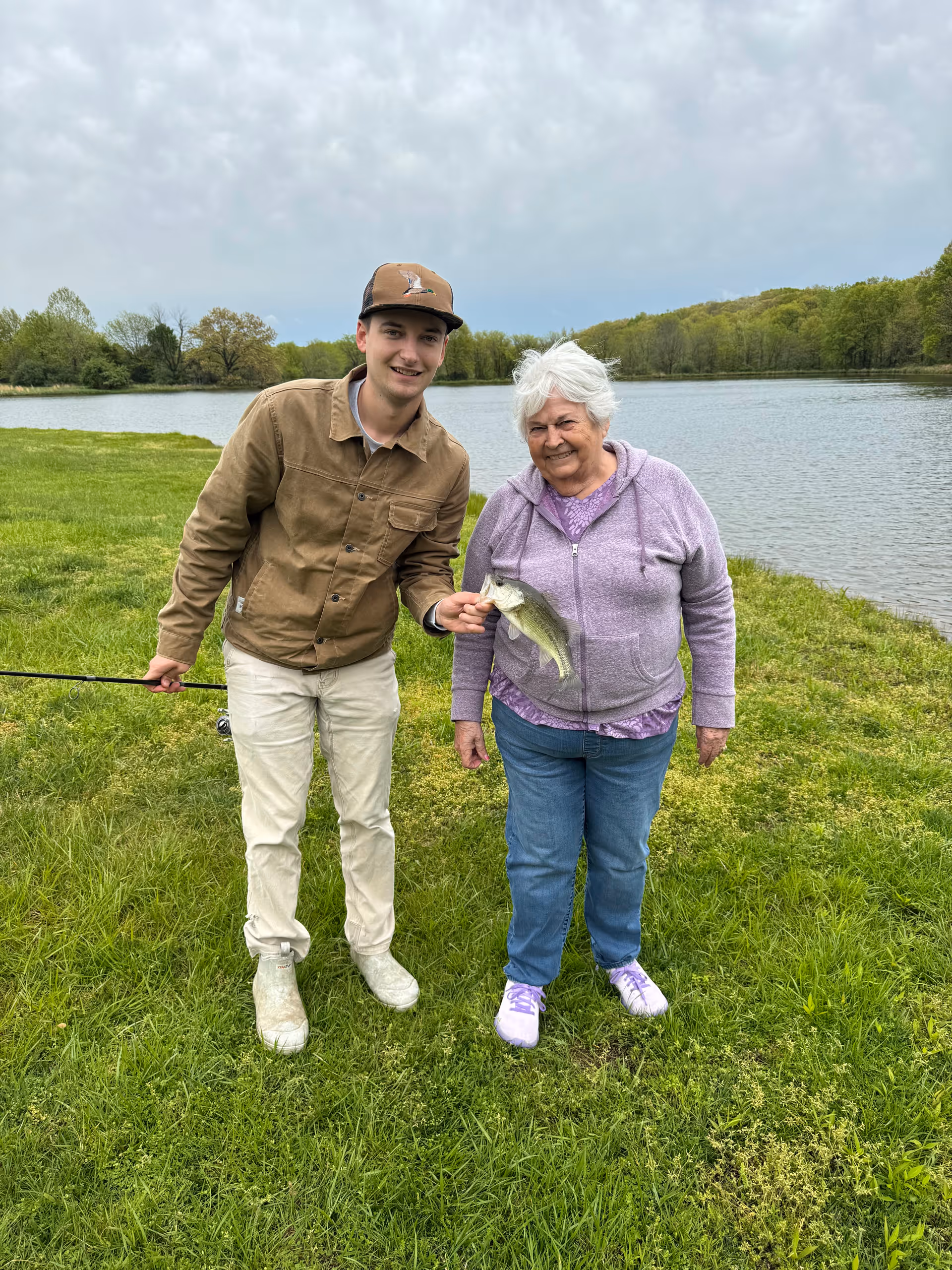 An elderly woman and a young man standing on grass near a lake. The young man is holding a fishing rod and the elderly woman is holding a fish they caught. Trees and water are visible in the background under a cloudy sky.