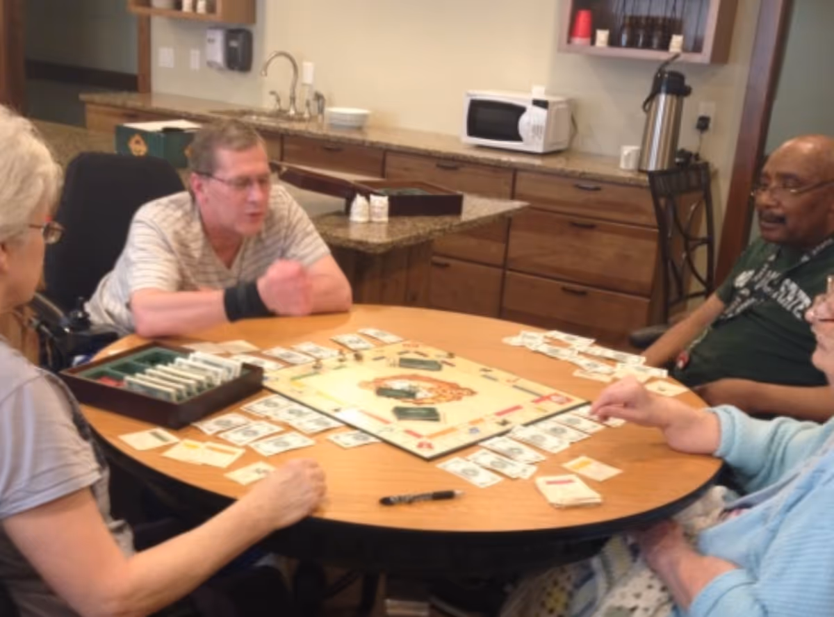 Four elderly individuals sitting around a round wooden table playing a board game resembling Monopoly in a room with wooden cabinets, a microwave, and a coffee dispenser in the background.