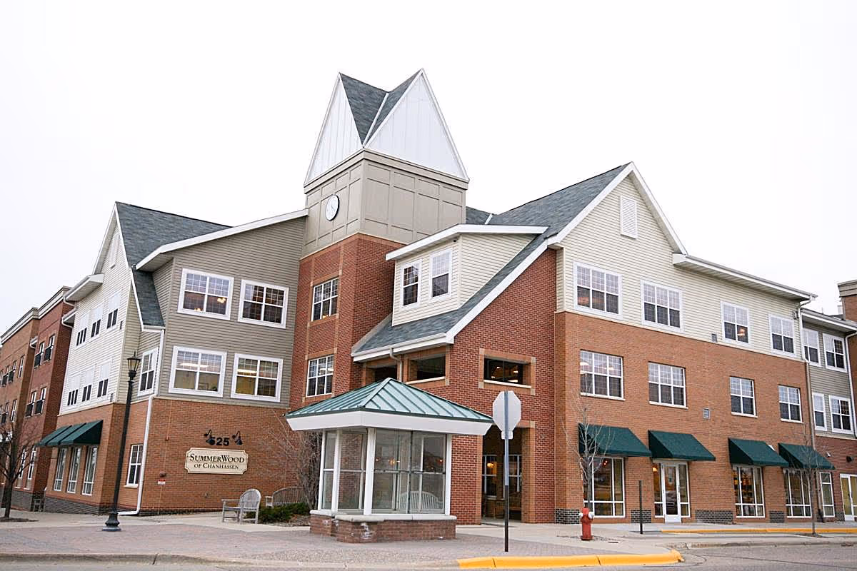 Exterior view of a multi-story senior living facility named SummerWood of Chanhassen, featuring a combination of brick and siding with multiple windows, green awnings, a clock tower, and a small glass-enclosed entrance area.
