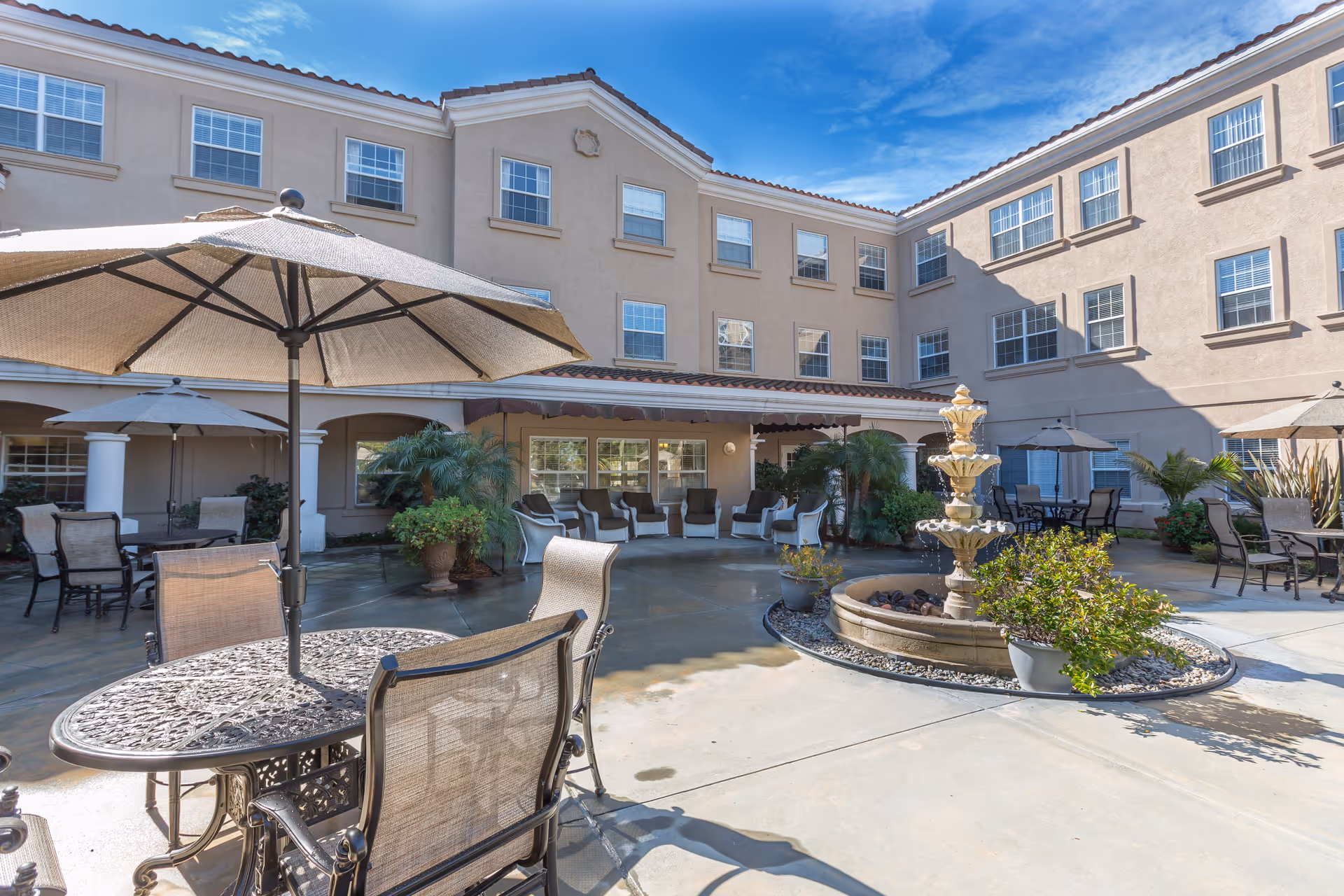Outdoor courtyard area of a senior living facility with multiple seating arrangements including metal tables and chairs with umbrellas. There is a three-tiered water fountain surrounded by plants in the center. The building surrounding the courtyard is three stories tall with many windows and a tiled roof under a clear blue sky.