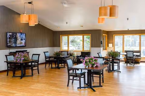 Spacious senior living dining area with wooden floors, tables, chairs, and potted flowers.