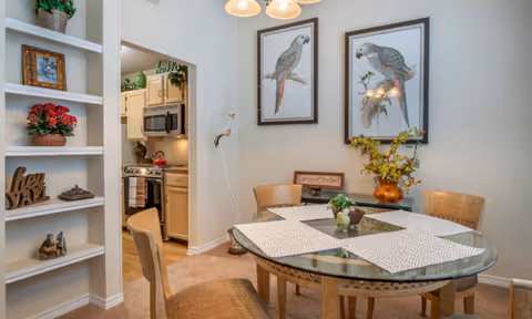 Dining area with a round glass table and four wicker chairs, bird artwork on the wall, and a view into a small kitchen.