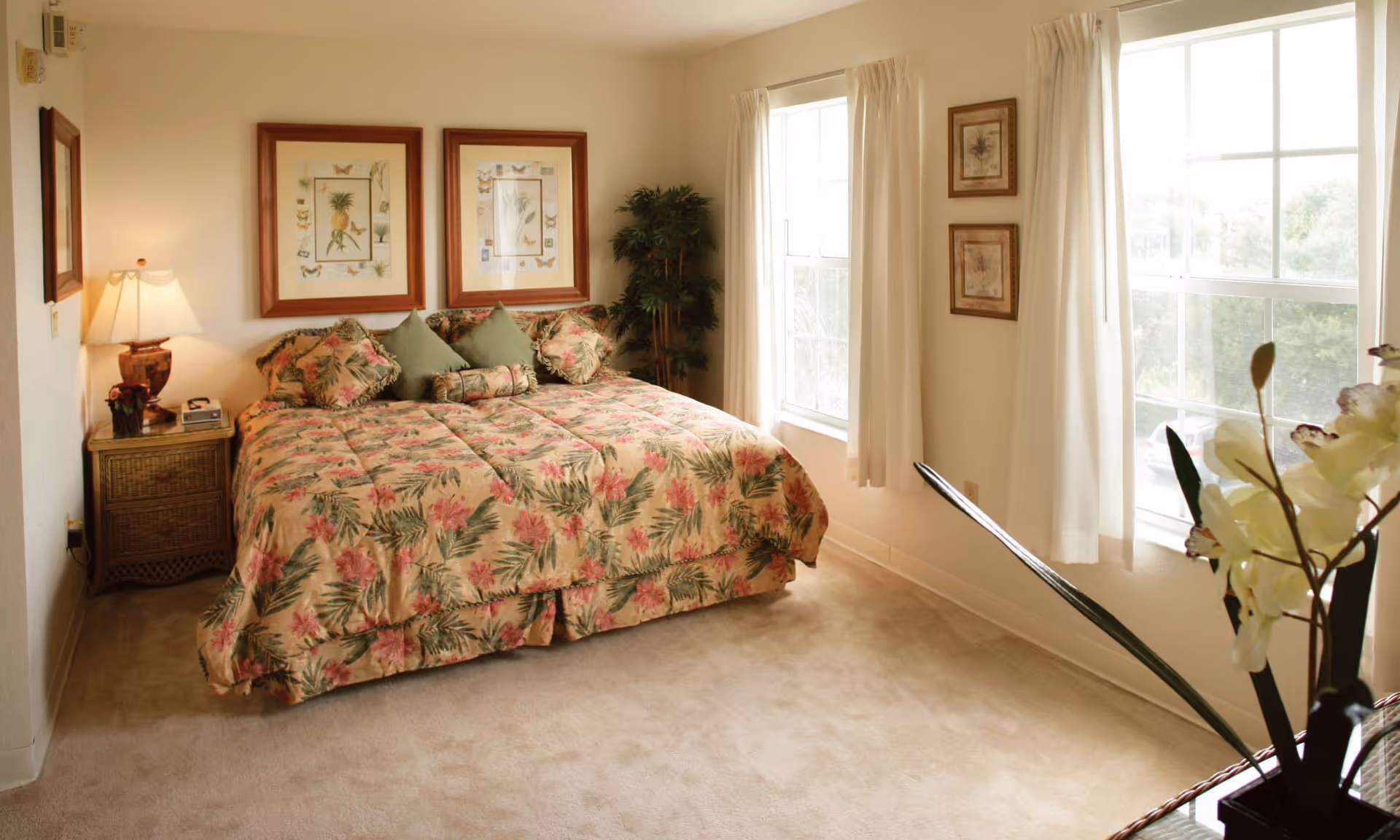 A cozy bedroom with a large bed covered in a floral patterned comforter and several pillows. There are two framed botanical prints hanging above the bed. A wooden nightstand with a lamp and a clock radio is beside the bed. The room has beige carpet, cream-colored walls, and two large windows with white curtains letting in natural light. A potted plant is placed in the corner near the windows, and a vase with white flowers is visible in the foreground.
