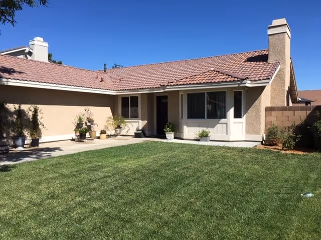 Single-story house with beige stucco walls and a red tile roof, featuring a well-maintained green lawn in the front yard and several potted plants near the entrance under a clear blue sky.