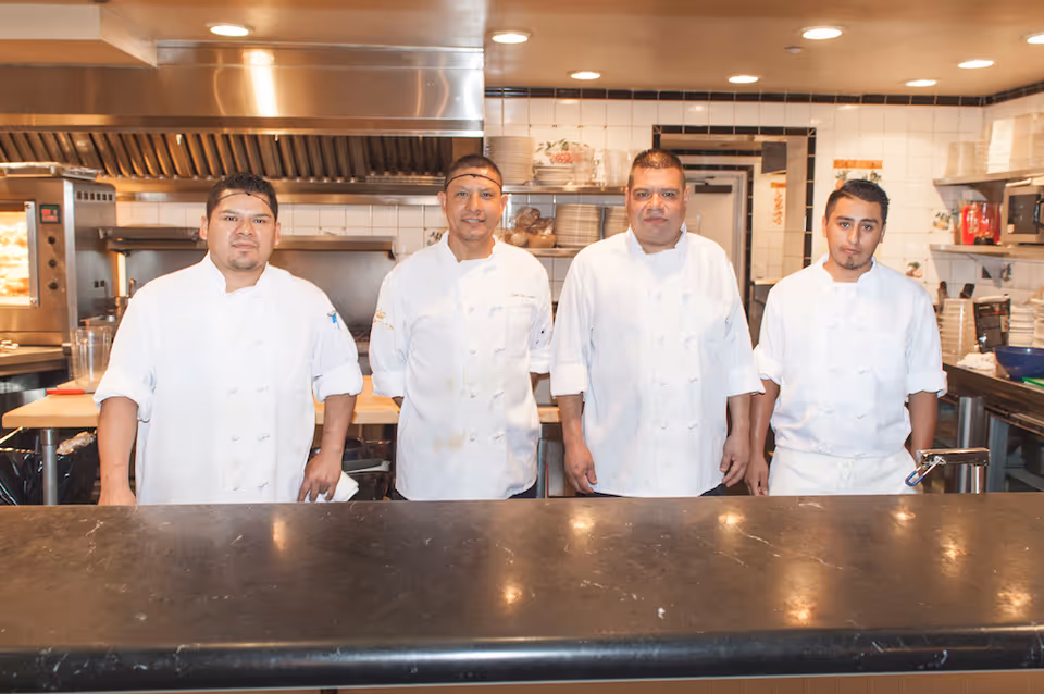 Four chefs wearing white uniforms standing behind a counter in a commercial kitchen with stainless steel appliances and shelves filled with dishes and kitchen supplies.