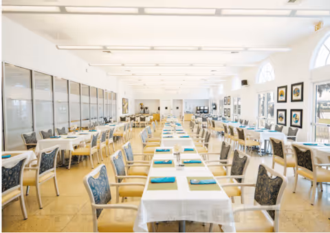 A bright and spacious dining room with long tables covered in white tablecloths, neatly arranged with turquoise napkins and place settings. The room features large windows with arched tops allowing natural light to fill the space, and walls adorned with framed artwork. Multiple rows of chairs with patterned upholstery are placed around the tables.