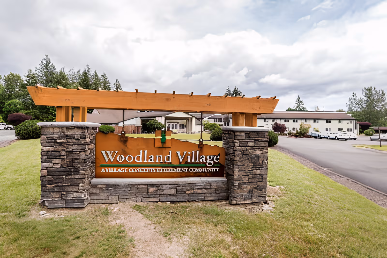 Stone and wood entrance sign reading Woodland Village with the facility building and driveway visible behind it.