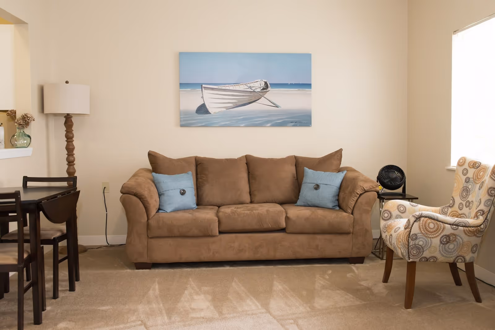 A cozy living room with a brown sofa adorned with two blue pillows, a patterned armchair, a wooden floor lamp, a small side table with a black fan, and a painting of a boat on the beach hanging on the wall above the sofa. There is also a small dining table with chairs visible on the left side.