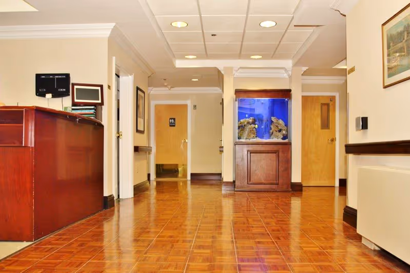 Interior hallway of a senior living facility with polished wooden floors, a wooden reception desk on the left, an aquarium with coral and fish in the center, and two closed wooden doors at the end of the hallway. The walls are light-colored with framed artwork and ceiling lights illuminating the space.