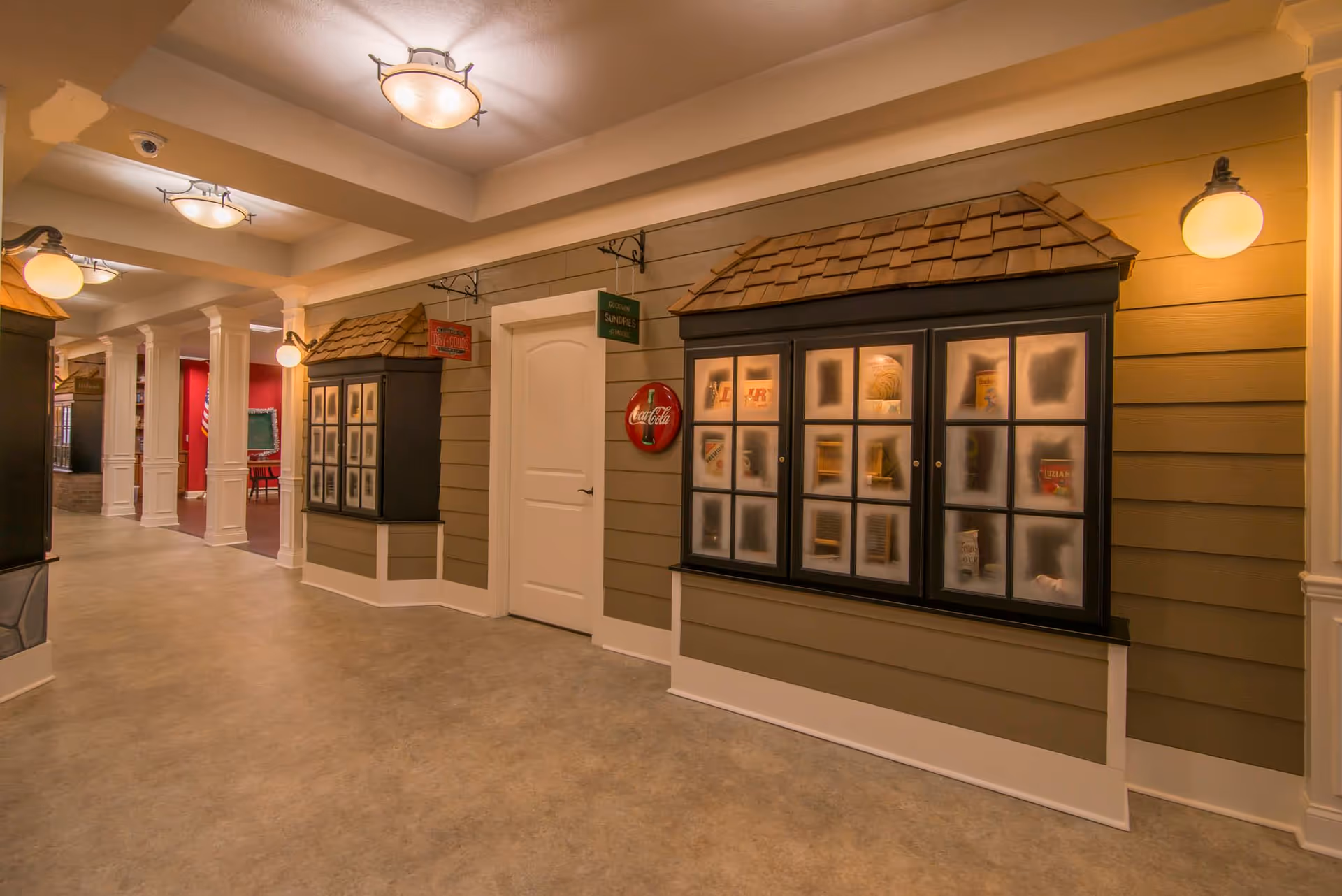 Indoor hallway with beige walls and white trim, featuring display cabinets with small shingled roofs and frosted glass showcasing various items. The hallway has ceiling lights and wall sconces, with a white door in the center and signs above it. In the background, there is a glimpse of a red room with an American flag and a chalkboard.