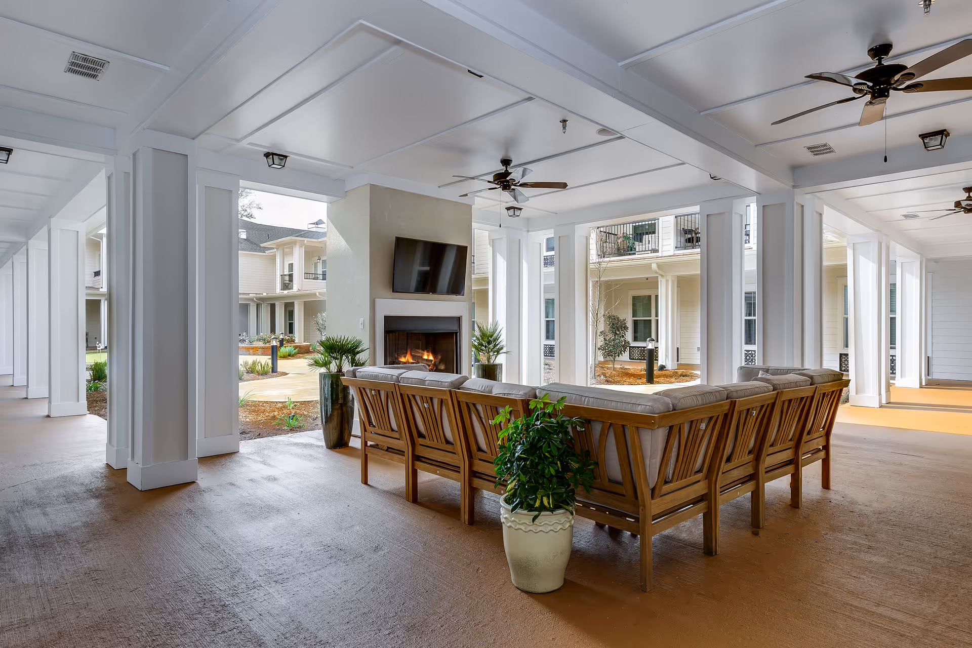 Covered courtyard lounge with a fireplace and wall-mounted TV, cushioned wooden seating, ceiling fans, and potted plants.