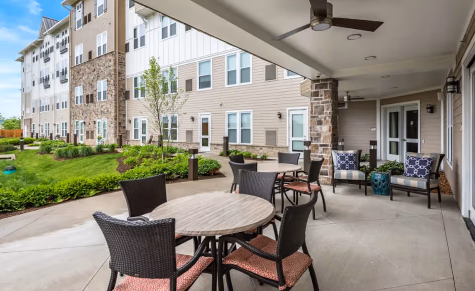 Outdoor covered patio area at Promenade Senior Living with round tables and chairs, cushioned seating, ceiling fan, and landscaped garden with grass and shrubs adjacent to the building.