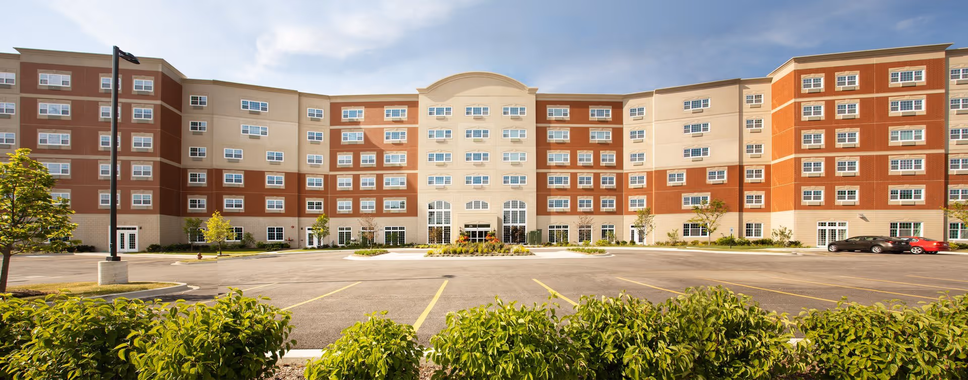 Wide exterior view of The Radcliff senior living facility showing a large multi-story building with numerous windows, a central entrance, a spacious parking lot in front, and landscaped greenery including bushes and small trees.