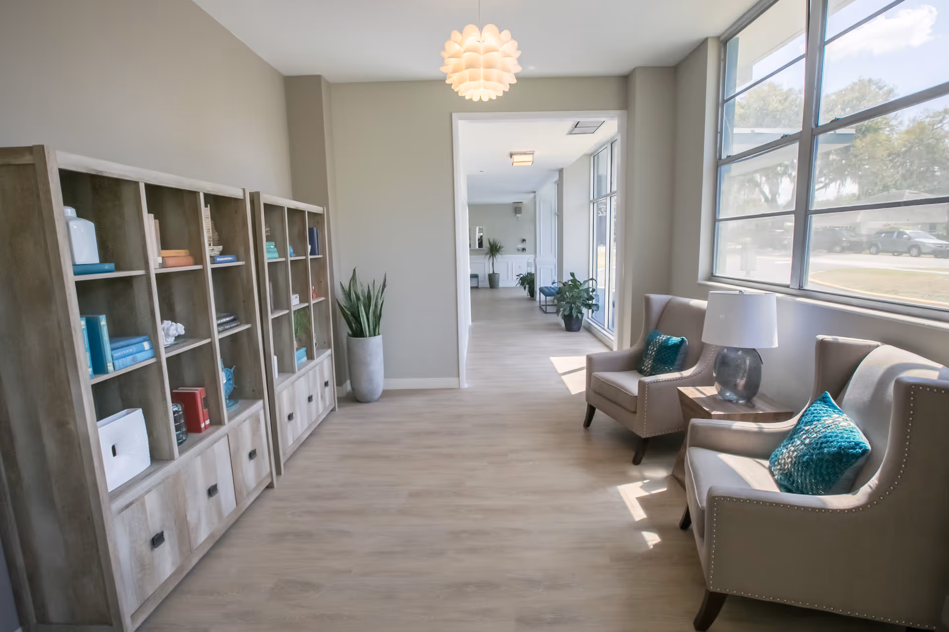 A bright and airy interior hallway in Lakeview Tower featuring large windows on the right side letting in natural light. Two beige armchairs with teal cushions and a small wooden side table with a lamp are placed near the windows. On the left side, there are wooden bookshelves with various decorative items and books. The hallway leads to another room with more plants and seating visible in the distance.