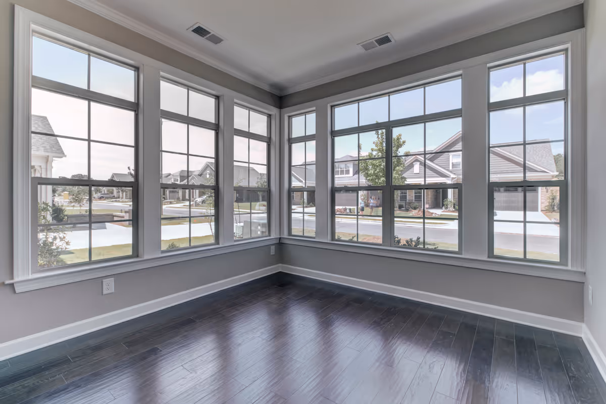 Empty room with large windows on two walls allowing natural light to fill the space, showing a view of a suburban neighborhood with houses and trees outside. The room has dark hardwood floors and light gray walls with white trim.