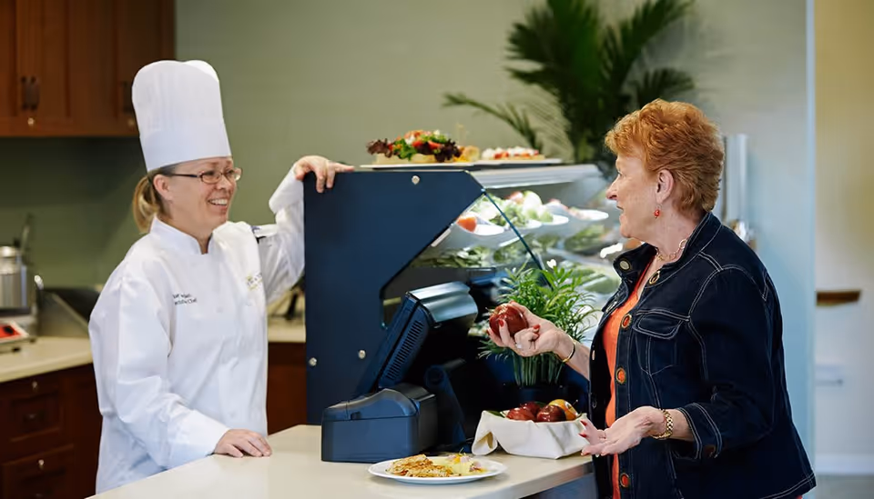 A chef wearing a white uniform and tall white chef hat smiles while talking to an elderly woman holding an apple at a food counter with plates of food and fruit bowls in the background.