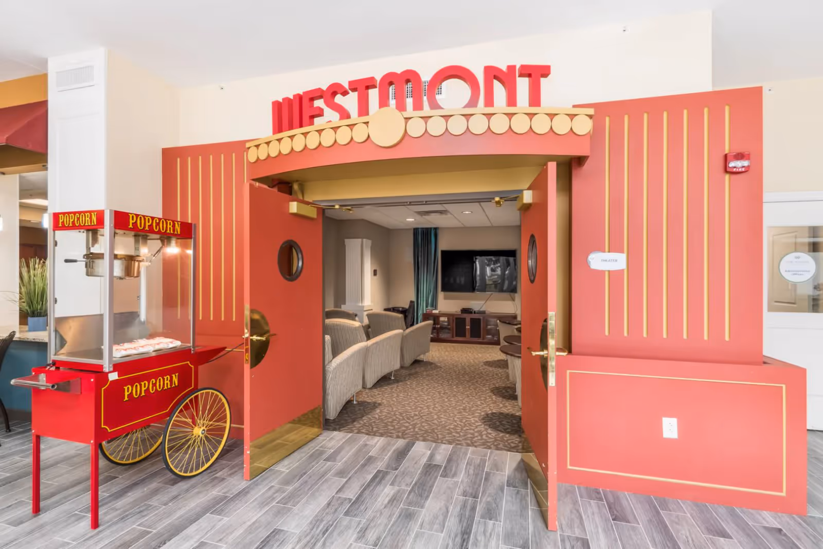 Red theater-style entrance labeled 'WESTMONT' with a popcorn cart and a seating area with a TV visible through the open doors.