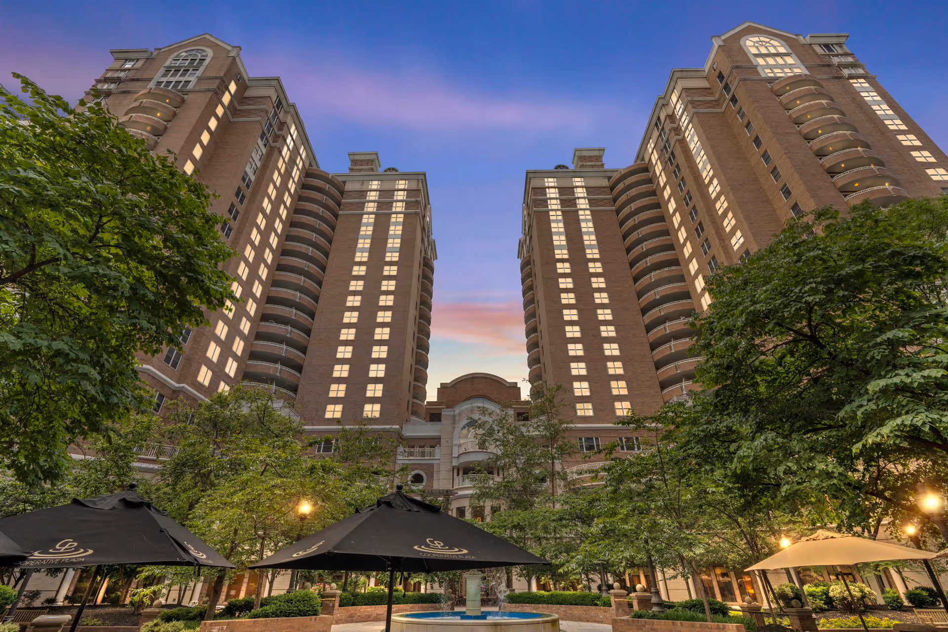 View of a tall, multi-story brick residential building with balconies, surrounded by trees and outdoor umbrellas over seating areas, under a twilight sky.