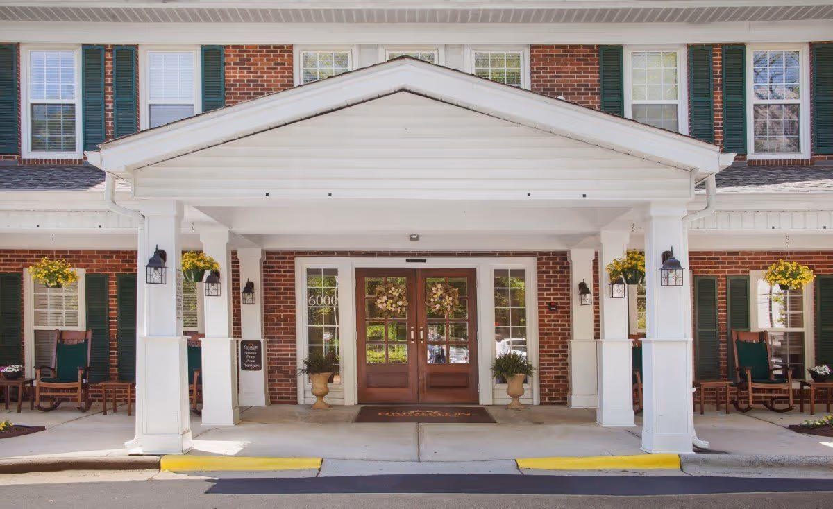 Covered entrance with white columns and double wooden doors set in a red brick facade, flanked by rocking chairs and hanging flower baskets.
